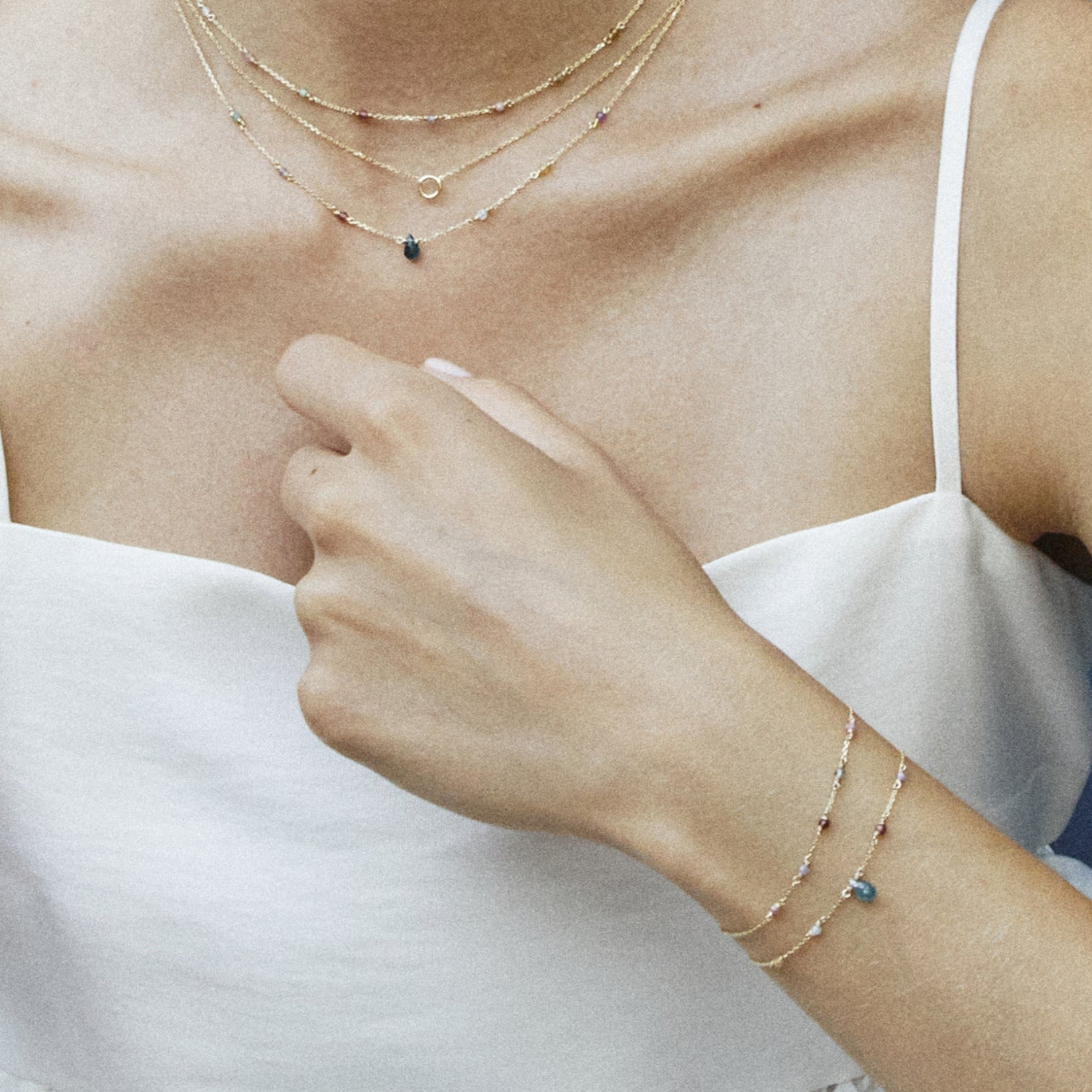 Drei zarte goldene Halsketten mit kleinen bunten Steinen und zwei goldene Armbänder mit blauen und violetten Steinen an einer Person in weißem ärmellosem Oberteil;