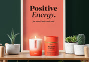 Aery Aromatherapy Kerze und Verpackung mit Aufschrift 'Positive Energy' auf einem Holztisch, umgeben von kleinen Sukkulenten in weißen Töpfen;
