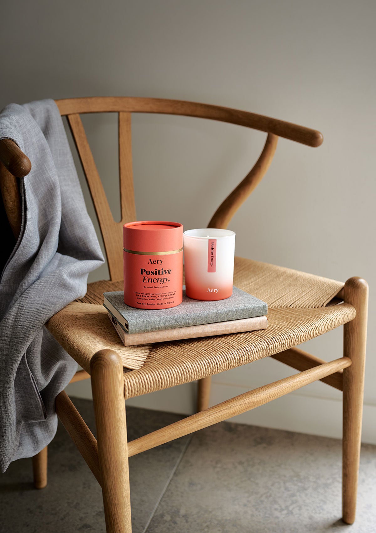 Aery Aromatherapy Kerze und Verpackung mit Aufschrift 'Positive Energy' auf einem Holztisch, auf zwei Büchern, auf einem Holzstuhl mit grauem Stoff über der Lehne;