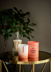 Aery Aromatherapie-Kerze 'Positive Energy' in weiß-orangem Glas mit passender orangefarbener Verpackung auf rundem goldfarbenem Tisch, Hintergrund mit grüner Pflanze und Glasbehälter mit Streichhölzern;