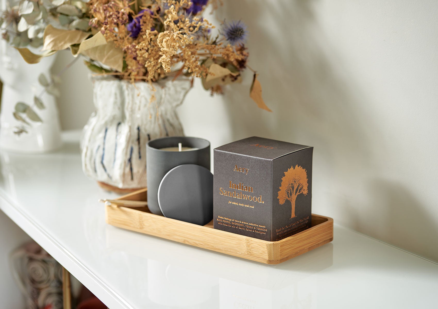 Aery Duftkerze 'Indian Sandalwood' in schwarzem Glas mit passendem Deckel und Verpackung auf einem Holztablett neben einer Vase mit getrockneten Blumen;