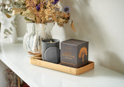 Aery Duftkerze 'Indian Sandalwood' in schwarzem Glas mit passendem Deckel und Verpackung auf einem Holztablett neben einer Vase mit getrockneten Blumen;