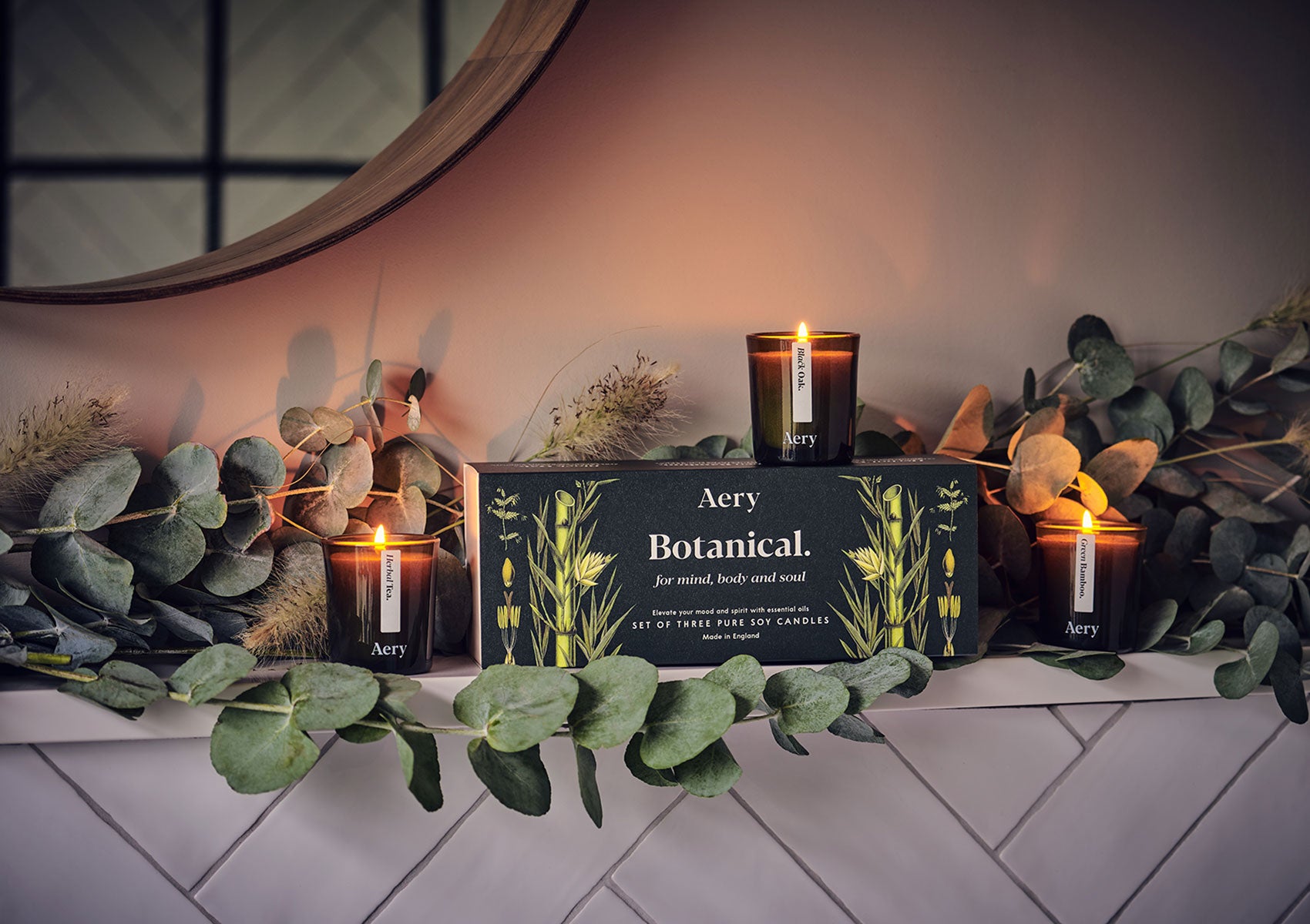 Drei brennende Aery Botanical Duftkerzen in grünen Gläsern neben schwarzer Geschenkbox mit Pflanzenmotiv auf weiß gekacheltem Regal mit Eukalyptuszweigen;