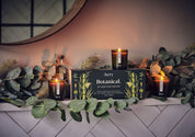 Drei brennende Aery Botanical Duftkerzen in grünen Gläsern neben schwarzer Geschenkbox mit Pflanzenmotiv auf weiß gekacheltem Regal mit Eukalyptuszweigen;