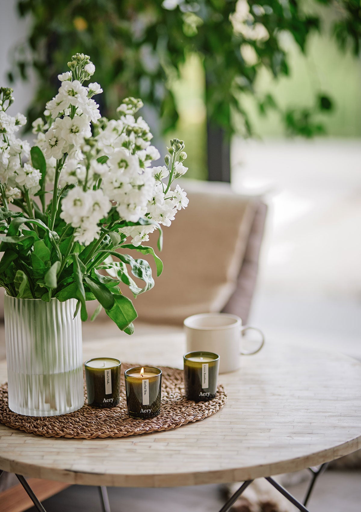 Weiße Blumen in Glasvase, drei grüne Aery Duftkerzen auf Tisch, weiße Tasse im Hintergrund;