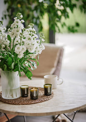 Weiße Blumen in Glasvase, drei grüne Aery Duftkerzen auf Tisch, weiße Tasse im Hintergrund;