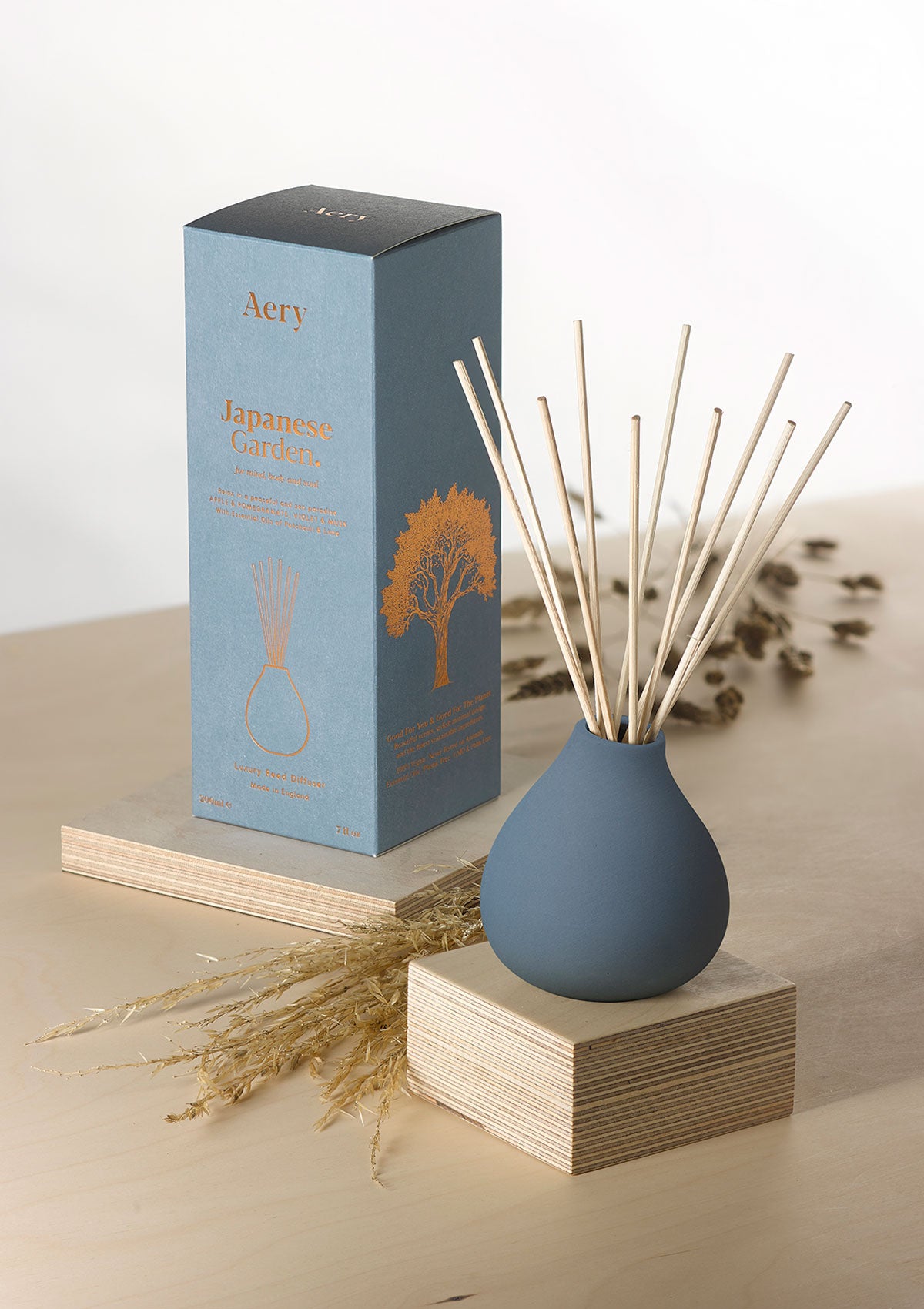 Aery Japanese Garden Diffuser in blauer, bauchiger Vase mit Holzstäbchen neben blauer Verpackung mit Baumillustration;