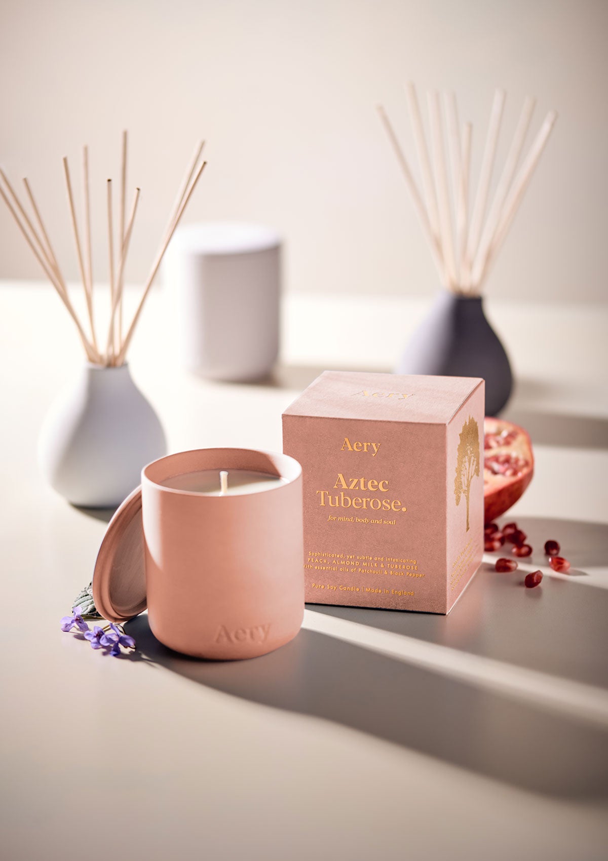 Aery Duftkerze 'Aztec Tuberose' in rosa Keramikbehälter mit passendem Deckel und Verpackung, daneben Granatapfelkerne und Duftstäbchen;