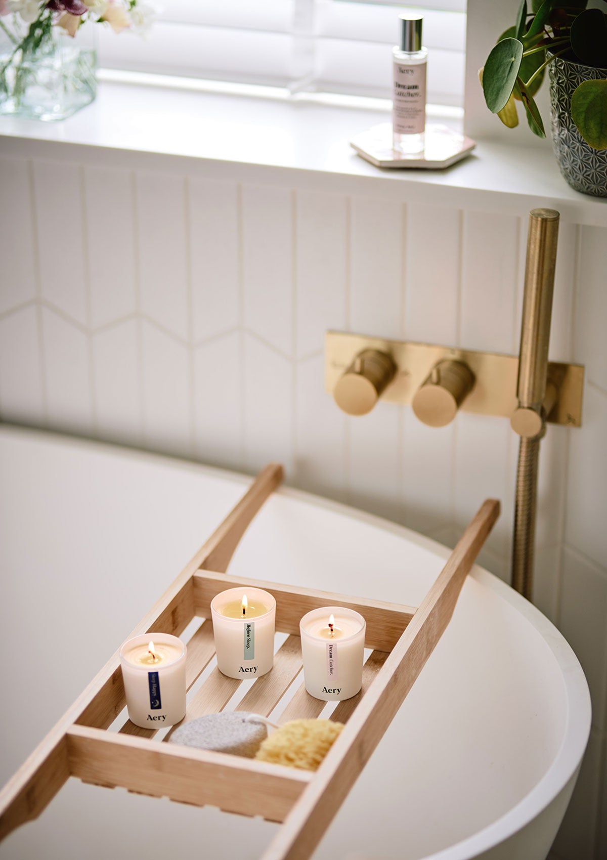 Drei weiße Aromatherapie-Kerzen von Aery mit den Düften 'Before Sleep', 'Sleep Happy' und 'Dream Catcher', die auf einer Badewannenablage aus Holz stehen;