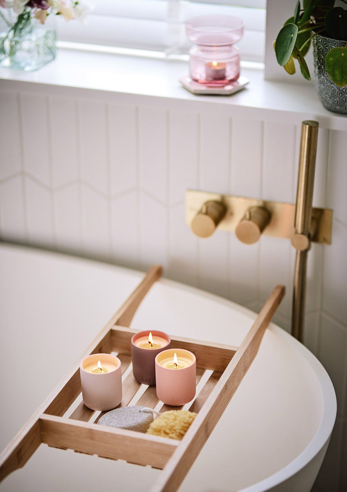 Badewanne mit Holzablage, darauf drei brennende Kerzen, Schwamm und Badeschwamm, goldfarbener Wasserhahn an weiß gefliester Wand;