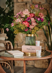 Bunter Blumenstrauß in grün geriffelter Glasvase auf rundem Glastisch mit zwei Kerzen und Geschenkset 'Happy Space' von Aery;
