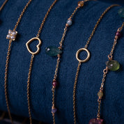 Fünf filigrane goldene Armbänder mit verschiedenen Anhängern: Perlenblume, Herz, grüne und rote Edelsteine, Kreis auf blauem Stoff;