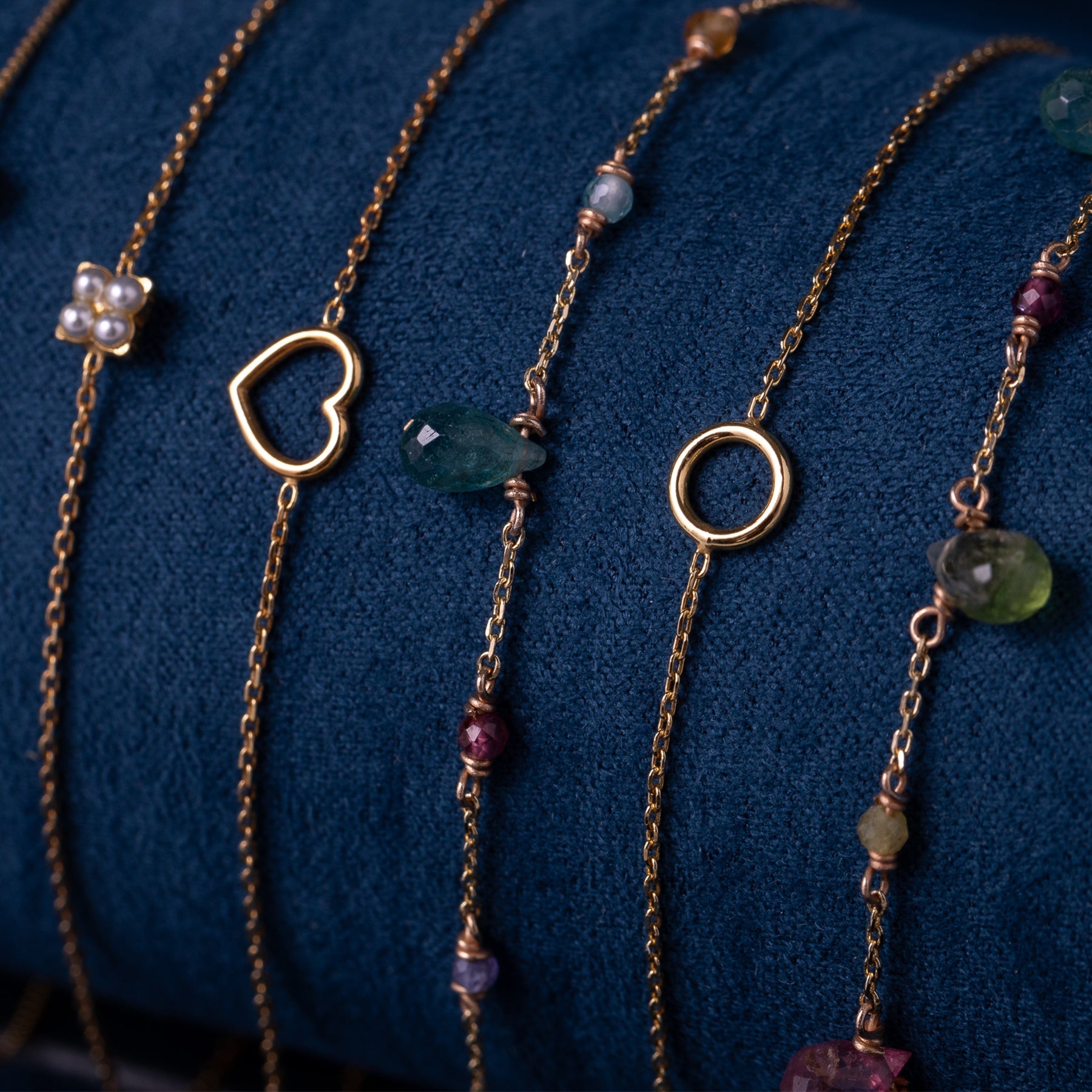 Fünf filigrane goldene Armbänder mit verschiedenen Anhängern: Perlenblume, Herz, grüne und rote Edelsteine, Kreis auf blauem Stoff;