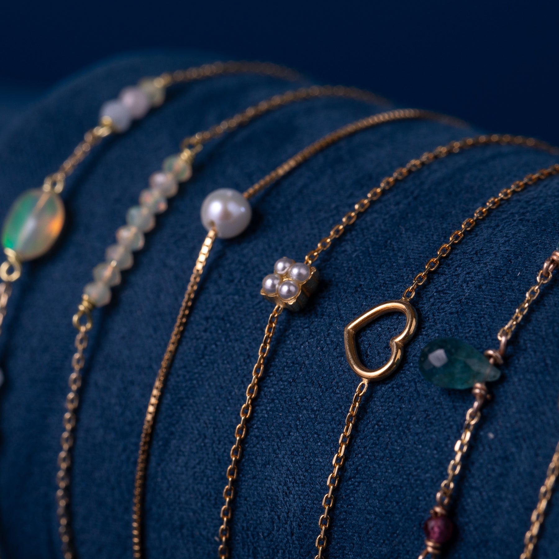 Feine goldfarbene Armbänder mit Perlen, Herzanhänger und Edelsteinen auf blauem Stoff;