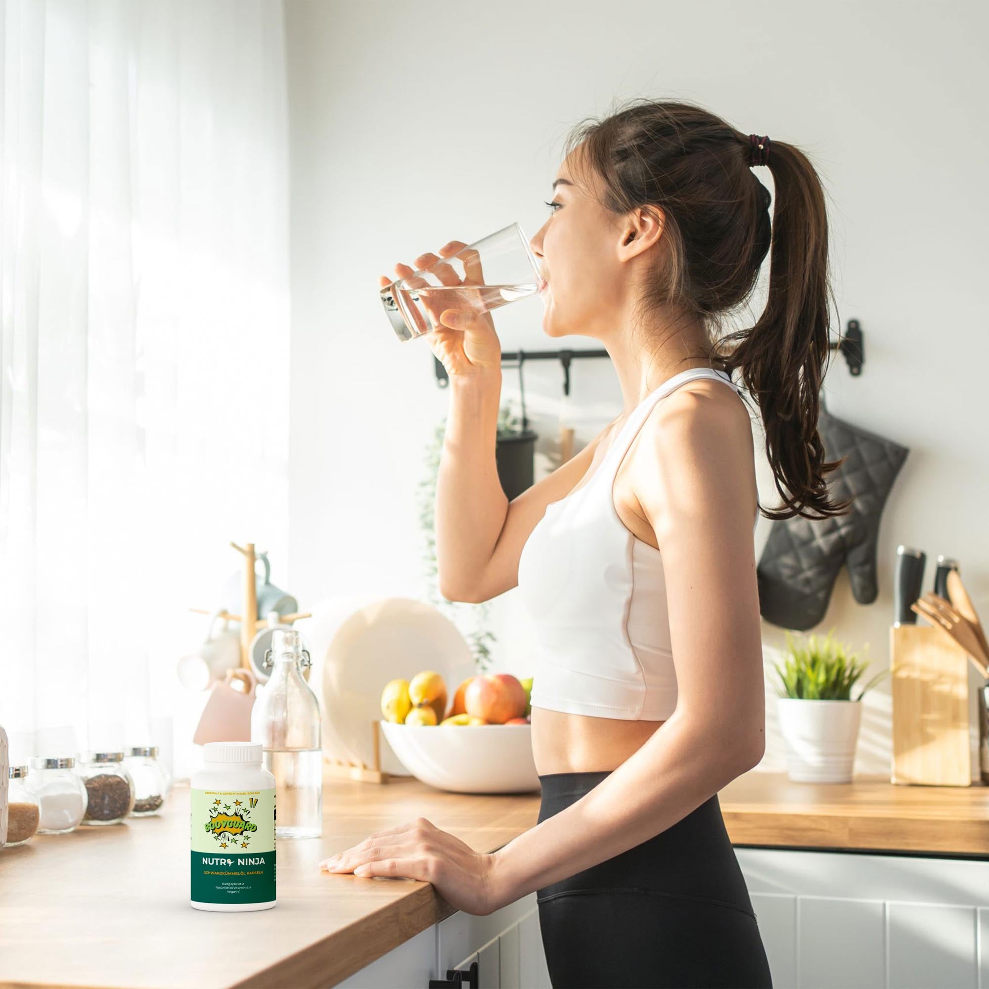 Frau mit Pferdeschwanz in Sportkleidung trinkt Wasser in heller Küche, steht am Tresen mit Nahrungsergänzungsmittel Nutri Ninja und Obstschale;