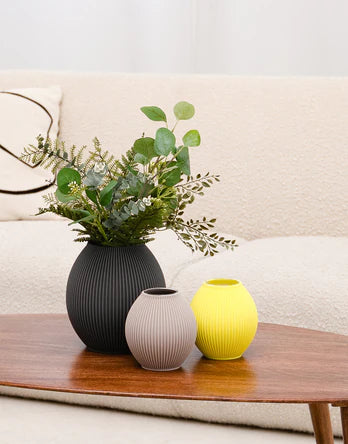 Drei kugelförmige Vasen mit gerippter Oberfläche stehen auf einem Holztisch, dahinter ein weißes Sofa;
in der größeren dunkelgrauen Vase sind grüne Zweige, daneben eine hellgraue und eine gelbe Vase;